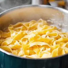 Szeroki, żółty makaron tagliatelle gotuje się w bulgoczącej wodzie w dużym, metalowym garnku.