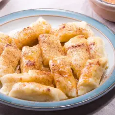 Talerz pełen najsmaczniejszych pierogów leniwych, polanych masłem i posypanych cynamonem. Obok widelec i miseczka z cukrem.