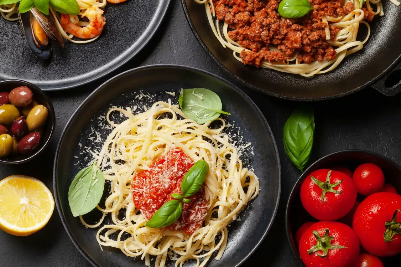 Pyszne spaghetti z sosem pomidorowym, posypane startym serem, ozdobione bazylią. Obok talerz z krewetkami i małżami, miseczka oliwek i świeże pomidory.