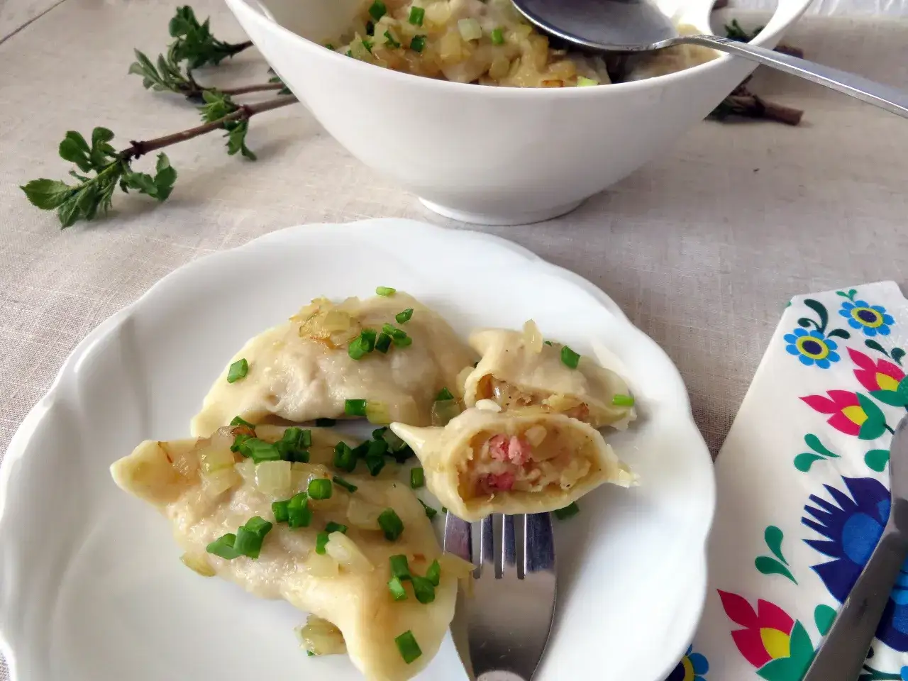 Pyszne pierogi chłopskie z cebulką i szczypiorkiem na białym talerzu. Jedne z nich są przekrojone, ukazując sycące nadzienie.